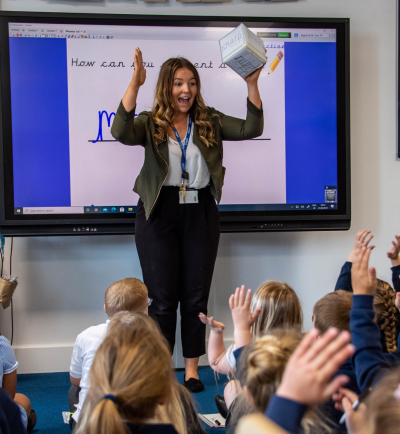 A teacher standing in front of her class of young children, raising her arms.