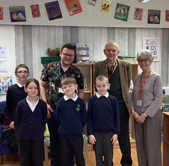 A group of children and two adults smiling. In the background is a white wall with cards on it.